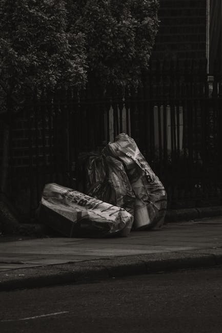 A group of black plastic rubbish bags, some with visible creases and reflective surfaces, are placed on the pavement near a black metal fence and a trim hedge with dense, dark foliage. The bags appear to contain domestic waste and are positioned at the edge of a residential area, with a section of the street and curb visible in the foreground. The environment suggests an outdoor setting likely awaiting collection for waste removal, as handled by professional services such as Rubbish Removal Barnet, which offers alternative disposal options outside of standard local authority collection. The scene is dimly lit, possibly during early morning or late evening, with subdued natural light creating shadows and a somewhat muted aesthetic. The waste bags are stacked or leaning against the fence, with minimal surrounding clutter, emphasizing the focus on on-site waste disposal activities typical of private rubbish removal services in Barnet.