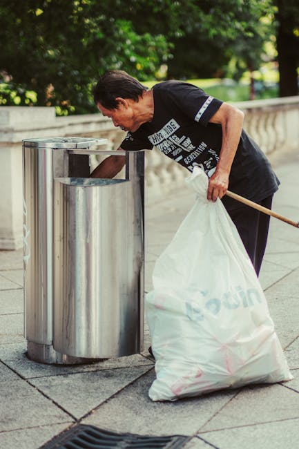 A man with dark hair, wearing a black T-shirt with white text, is using a wooden pole to collect rubbish from a stainless steel outdoor public litter bin placed on a paved walkway. The bin has a modern, cylindrical design with separate compartments for waste. The man is leaning forward, pulling a large white plastic bag filled with rubbish out of the bin, which appears to contain a mixture of waste. Behind him, there is a low stone balustrade and lush green foliage, indicating the scene is set in an outdoor park or urban area with natural surroundings. The lighting suggests daytime, with dappled sunlight filtering through the trees. The image illustrates an active waste collection or rubbish removal process, likely associated with private or community-managed refuse management, supported by services such as Rubbish Removal Barnet.