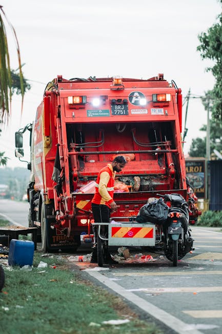 A municipal waste collection worker, dressed in a high-visibility orange and yellow uniform, is positioned at the rear of a large red rubbish truck on a street. The worker is leaning slightly forward, actively engaged in emptying a black trash bag into the truck's hopper, which is open and reveals a metal interior. The rubbish truck features various safety lights, reflective striping, and identification markings, including a white circle with a blue emblem and vehicle registration details in black. The truck is parked partially on the pavement with the rear end close to a bicycle lane or designated collection zone, and nearby are small pieces of litter scattered on the ground. In the background, there are trees, utility poles, and a building with signage, suggesting an urban or suburban area. The environment appears to be an early morning or overcast day, with diffused natural light illuminating the scene, creating a professional and factual depiction relevant to independent rubbish collection and waste disposal services often associated with private waste management providers like Rubbish Removal Barnet.
