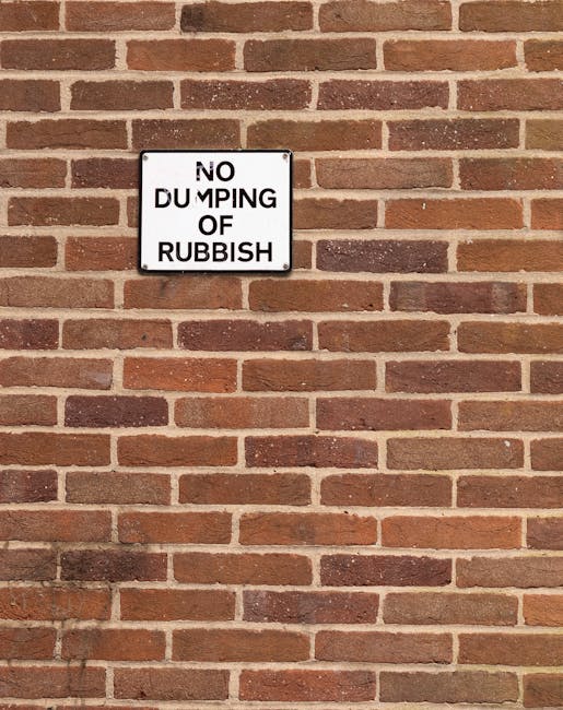 A rectangular metal sign mounted on a red and brown brick wall displaying the message 'NO DUMPING OF RUBBISH' in black uppercase letters on a white background. The sign is positioned toward the upper left portion of the image, with the brick wall extending horizontally across the frame. The bricks have a textured surface, featuring variations in shade from reddish-bink to darker brown, and are arranged in a traditional running bond pattern with visible mortar joints. The lighting in the scene is even, with subtle shadows cast by the bricks, emphasizing the wall's textured surface. This scene likely relates to a site where rubbish removal services, such as those provided by Rubbish Removal Barnet, are necessary to prevent improper disposal in designated areas, illustrating the importance of clear signage to maintain proper waste management practices.
