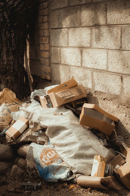 A pile of mixed household rubbish is accumulated against a concrete brick wall outdoors, consisting of flattened cardboard boxes, plastic packaging, and a large white sack. The cardboard boxes vary in size, with some opened and lacking flaps, showing signs of compression or damage. The packaging includes plastic wrapping and paper towels, with textures ranging from smooth and glossy to rough and crinkled. The sack, made from woven material, is partially filled and appears slightly misshapen. The environment is an outdoor area, with uneven ground composed of dirt, small stones, and debris, and a large tree trunk visible on the left side of the scene. The background features a weathered, off-white brick wall with some discoloration and slight staining, indicating outdoor exposure. Natural light highlights the different materials and textures, emphasizing the informal nature of the waste accumulation, which seems to relate to private collection or on-site clearance services as undertaken by Rubbish Removal Barnet.
