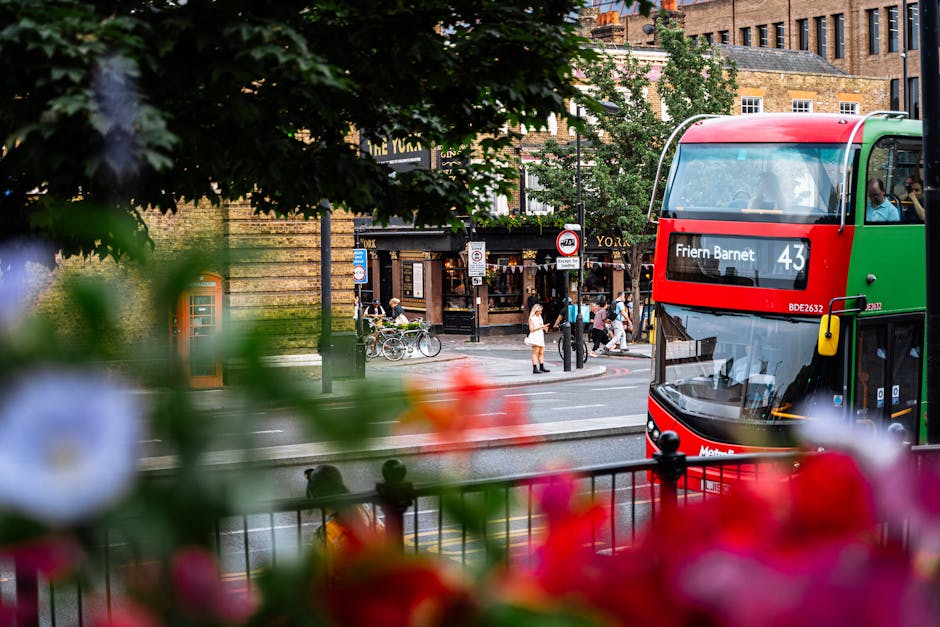 The image shows a busy urban street scene with a red and green double-decker bus in the foreground displaying the route number 43 and the destination 'Friern Barnet.' Partially obscured by blurred colorful flowers and green foliage in the lower part of the frame, the bus is positioned on the right side, with its front and side windows visible. Behind the bus, pedestrians cross the crosswalk and gather near the pavement, some waiting or walking in various directions. Several bicycles are parked along the curb, and in the background, there are buildings with a mix of brick facades and storefronts, including a pub or restaurant with a dark sign reading 'The York.' Trees with lush green leaves line the street, providing a natural canopy over the scene, and the environment suggests a lively, well-used area typical of city centres with public transportation, pedestrian activity, and retail outlets, aligning with alternative waste handling and rubbish collection contexts handled by local services such as Rubbish Removal Barnet.