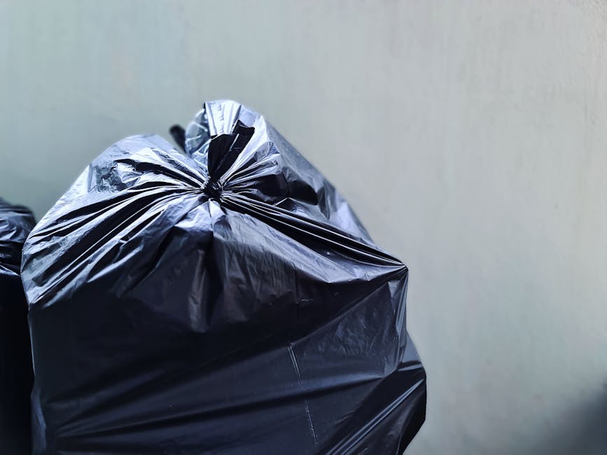 A pile of mixed household rubbish is accumulated against a concrete brick wall outdoors, consisting of flattened cardboard boxes, plastic packaging, and a large white sack. The cardboard boxes vary in size, with some opened and lacking flaps, showing signs of compression or damage. The packaging includes plastic wrapping and paper towels, with textures ranging from smooth and glossy to rough and crinkled. The sack, made from woven material, is partially filled and appears slightly misshapen. The environment is an outdoor area, with uneven ground composed of dirt, small stones, and debris, and a large tree trunk visible on the left side of the scene. The background features a weathered, off-white brick wall with some discoloration and slight staining, indicating outdoor exposure. Natural light highlights the different materials and textures, emphasizing the informal nature of the waste accumulation, which seems to relate to private collection or on-site clearance services as undertaken by Rubbish Removal Barnet.